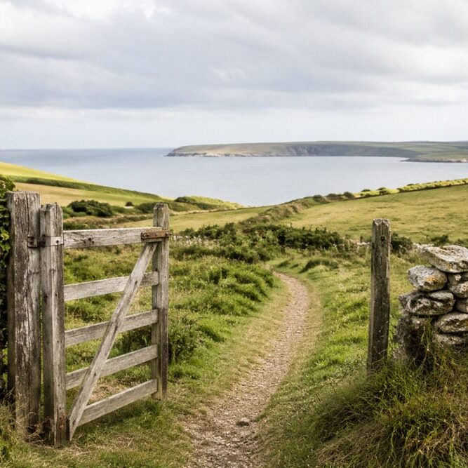 Open wooden gate leading along a coastal path towards the sea.
